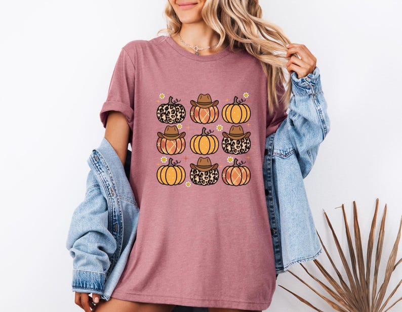 Puede incluir: Una camiseta malva con un estampado gr&aacute;fico de nueve calabazas con sombreros de vaquero. Las calabazas son de color naranja y marr&oacute;n con un estampado de leopardo.
