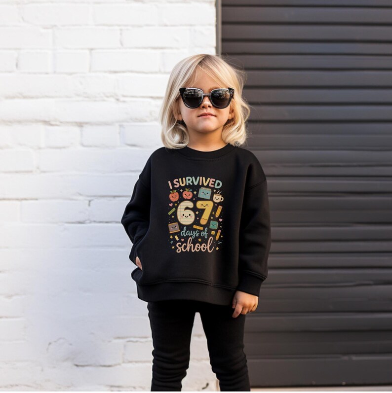 May include: A child wearing a black sweatshirt with the text "I Survived 67 days of school." The sweatshirt has colorful graphics of school-related items. The child is also wearing black pants and sunglasses.