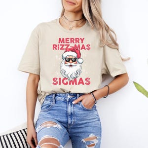 May include: A beige t-shirt with a graphic of Santa Claus wearing sunglasses and a red hat. The text "Merry Rizzmas Sigmas" is printed on the shirt.