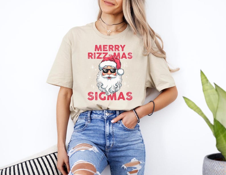May include: A beige t-shirt with a graphic of Santa Claus wearing sunglasses and a red hat. The text "Merry Rizzmas Sigmas" is printed on the shirt.