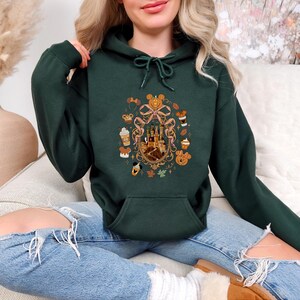 May include: Dark green hoodie with a front pocket and drawstring. The hoodie features a colorful graphic of a castle, pumpkins, and fall-themed treats. The design is surrounded by a decorative ribbon. The hoodie is made of a soft material.