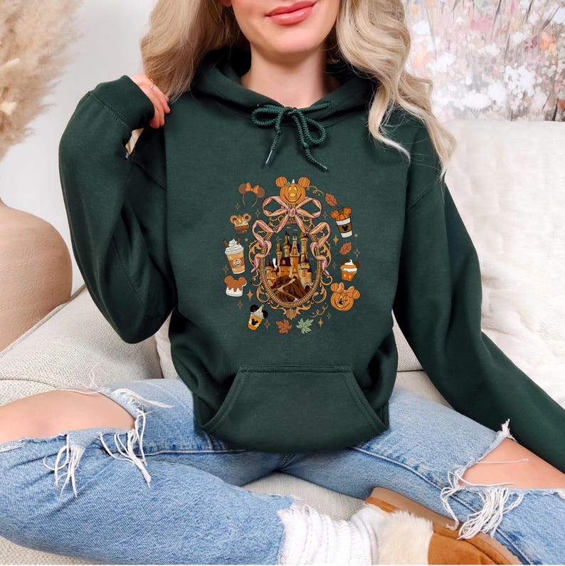 May include: Dark green hoodie with a front pocket and drawstring. The hoodie features a colorful graphic of a castle, pumpkins, and fall-themed treats. The design is surrounded by a decorative ribbon. The hoodie is made of a soft material.