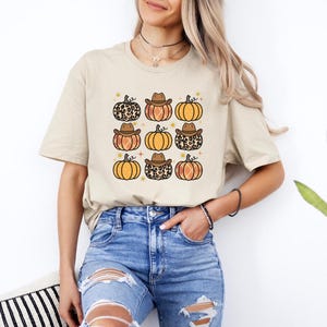 Puede incluir: Una camiseta beige con un patr&oacute;n de calabazas que llevan sombreros de vaquero. Las calabazas son de color naranja, marr&oacute;n y tienen un dise&ntilde;o de estampado de leopardo.