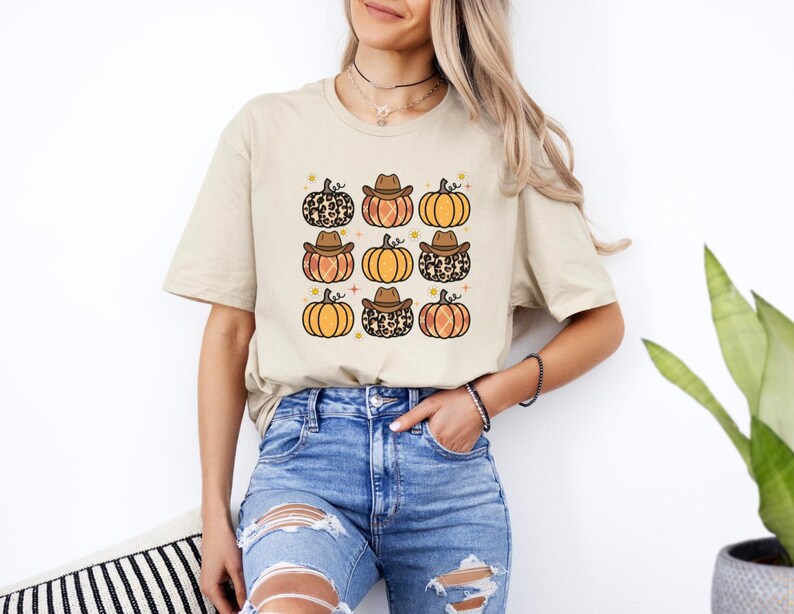 Puede incluir: Una camiseta beige con un patr&oacute;n de calabazas que llevan sombreros de vaquero. Las calabazas son de color naranja, marr&oacute;n y tienen un dise&ntilde;o de estampado de leopardo.