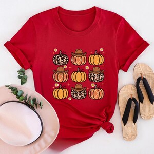 Puede incluir: Camiseta roja de manga corta con un estampado gr&aacute;fico de calabazas con sombreros de vaquero y estampado de leopardo. Las calabazas son de color naranja con tallos marrones y est&aacute;n rodeadas de peque&ntilde;as flores blancas.