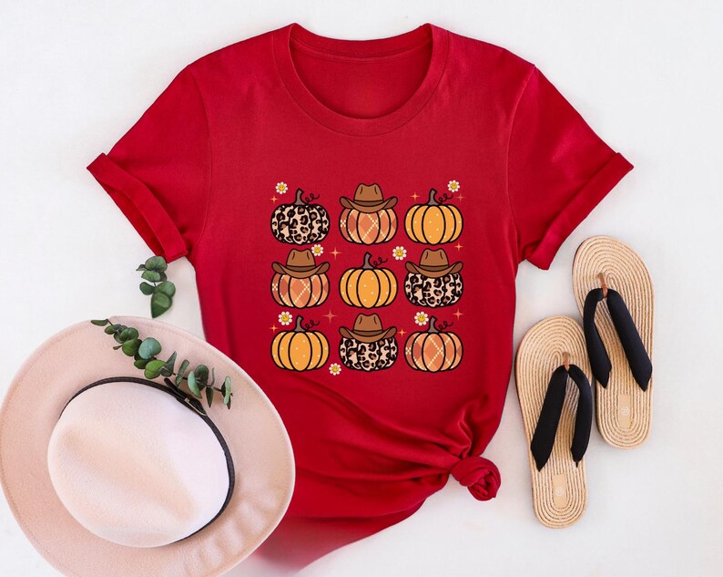 Puede incluir: Camiseta roja de manga corta con un estampado gr&aacute;fico de calabazas con sombreros de vaquero y estampado de leopardo. Las calabazas son de color naranja con tallos marrones y est&aacute;n rodeadas de peque&ntilde;as flores blancas.