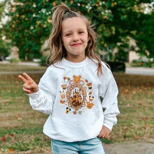 May include: A white long-sleeved sweatshirt featuring a colorful graphic of a castle, pumpkins, Mickey Mouse ears, and fall-themed elements. The design is centered on the front of the shirt.
