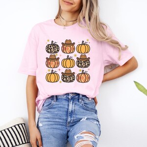 Puede incluir: Una camiseta rosa con un gr&aacute;fico de nueve calabazas con sombreros de vaquero. Algunas de las calabazas tienen un patr&oacute;n de estampado de leopardo.