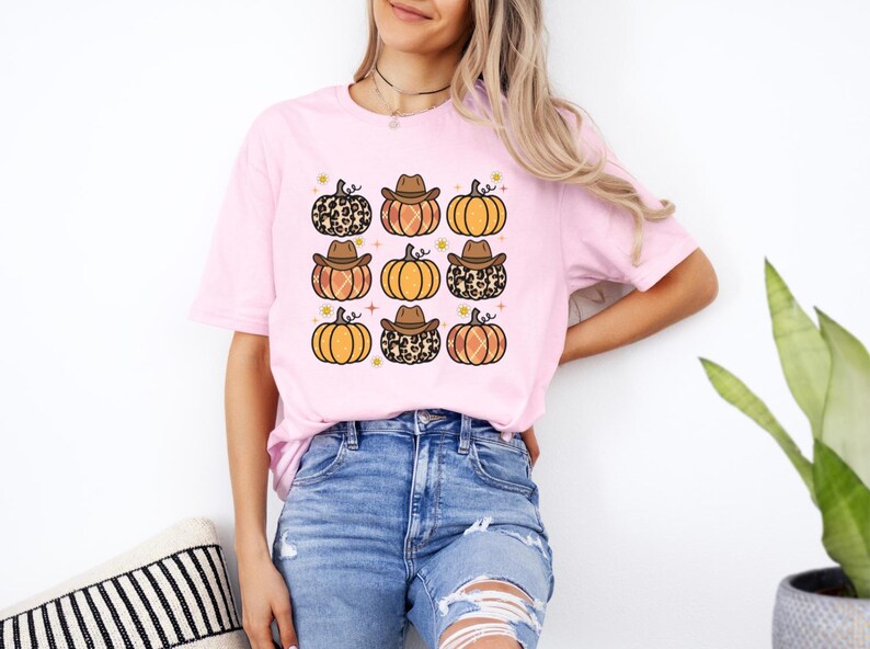 Puede incluir: Una camiseta rosa con un gr&aacute;fico de nueve calabazas con sombreros de vaquero. Algunas de las calabazas tienen un patr&oacute;n de estampado de leopardo.