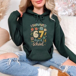 May include: A dark green hooded sweatshirt with the text "I Survived 67 days of school" in a colorful, playful font. The design includes illustrations of apples, books, and coffee cups. The sweatshirt has a front pocket and a drawstring hood.