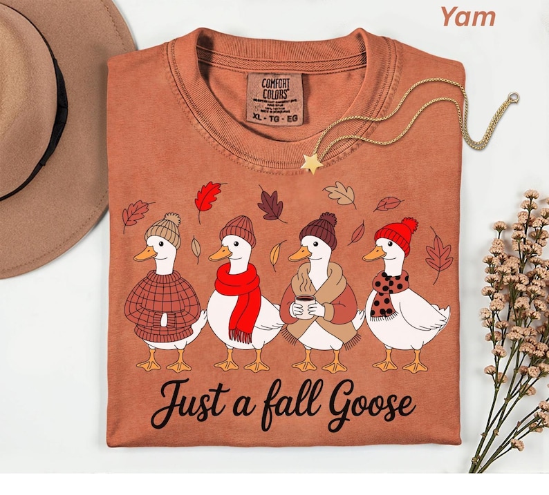 May include: A yam-colored t-shirt featuring a whimsical illustration of four geese dressed in fall attire. The geese are wearing hats, scarves, and sweaters, with the text "Just a fall Goose" below. A gold necklace with a star pendant is draped across the shirt.