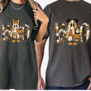 May include: Two dark gray t-shirts with a Halloween-themed design. The shirts feature the word "BOO" with a ghost character and a jack-o'-lantern. One ghost has a bow, the other has Mickey Mouse ears. The shirts are made of a soft material.