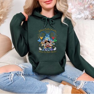 Peut inclure: Sweat-shirt &agrave; capuche vert fonc&eacute; avec un motif Disney Family Vacation. Le graphisme comprend des personnages Disney, un ch&acirc;teau, des feux d'artifice et l'ann&eacute;e 2026. Le texte "Where The Magic Begins" est &eacute;galement pr&eacute;sent. Le sweat-shirt a une poche avant et une capuche &agrave; cordon.