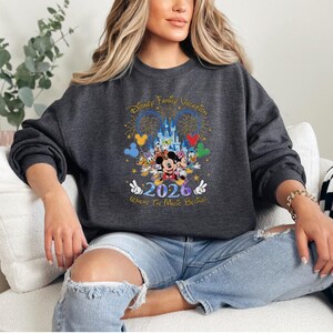 Peut inclure: Sweat-shirt gris fonc&eacute; avec un graphisme color&eacute; repr&eacute;sentant des personnages Disney, un ch&acirc;teau, des feux d'artifice et le texte "Disney Family Vacation 2026 Where The Magic Begins!"