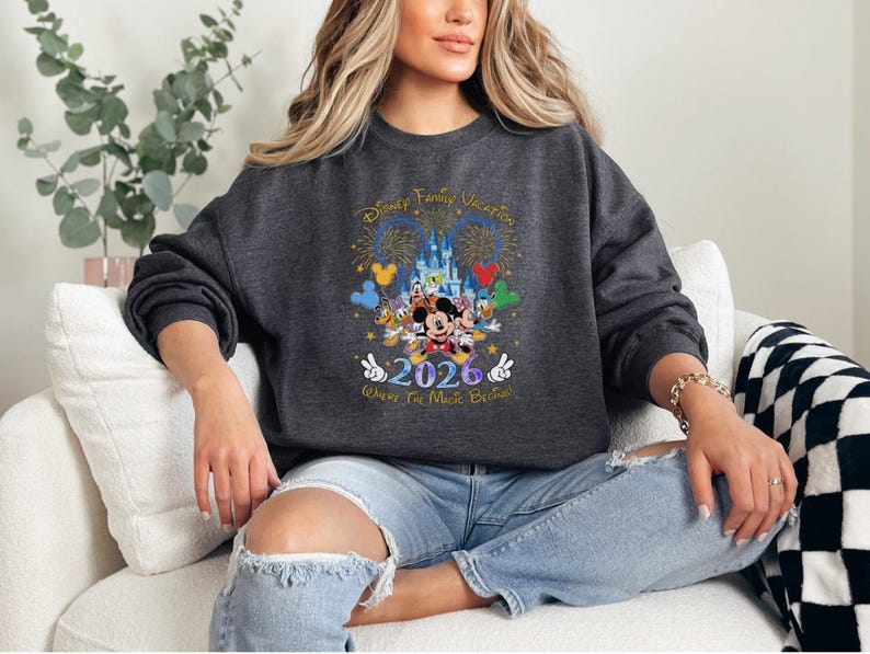 Peut inclure: Sweat-shirt gris fonc&eacute; avec un graphisme color&eacute; repr&eacute;sentant des personnages Disney, un ch&acirc;teau, des feux d'artifice et le texte "Disney Family Vacation 2026 Where The Magic Begins!"