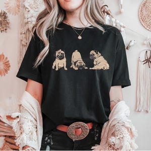 May include: Black t-shirt featuring a graphic of three pugs in different poses. The pugs are depicted in shades of tan and brown. The shirt is a classic crew neck style.