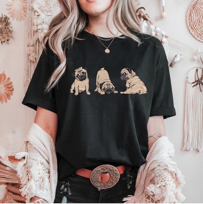 May include: Black t-shirt featuring a graphic of three pugs in different poses. The pugs are depicted in shades of tan and brown. The shirt is a classic crew neck style.