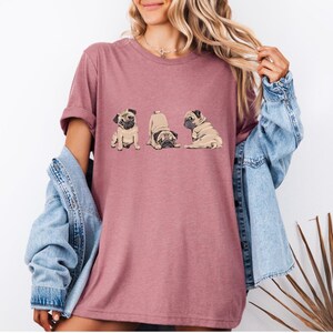 May include: A mauve-colored t-shirt featuring a graphic of three pugs in different poses. The pugs are in shades of brown and tan. The shirt is being worn by a person with long blonde hair and a denim jacket.