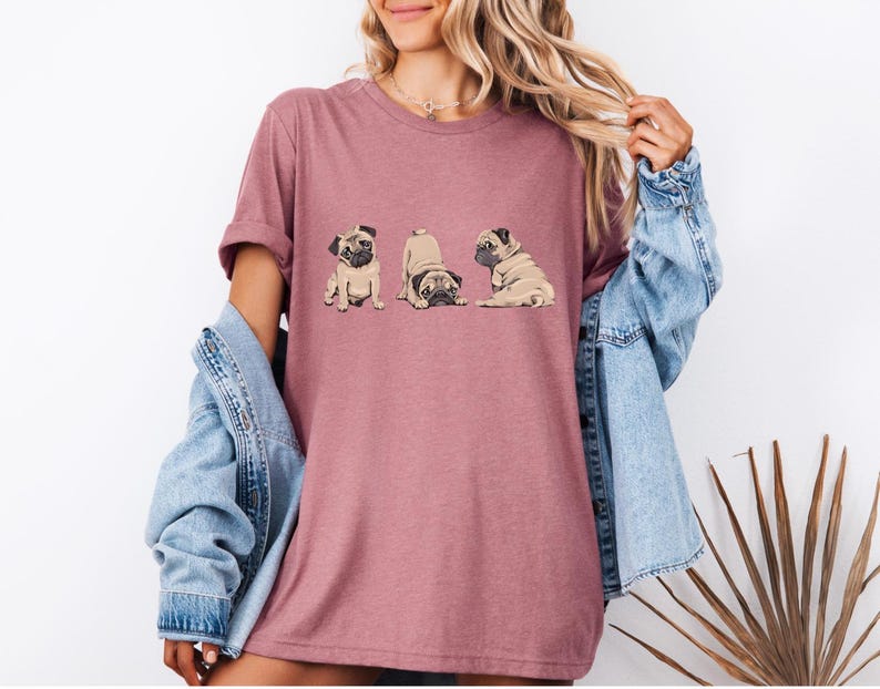 May include: A mauve-colored t-shirt featuring a graphic of three pugs in different poses. The pugs are in shades of brown and tan. The shirt is being worn by a person with long blonde hair and a denim jacket.
