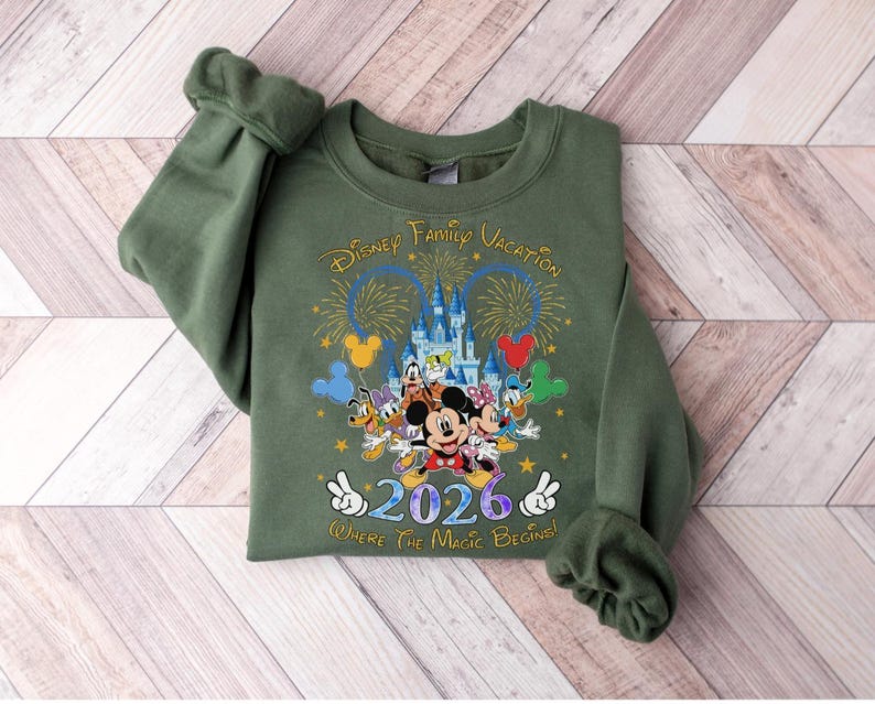 Peut inclure: Un sweat-shirt vert olive avec un graphisme color&eacute; de personnages Disney, un ch&acirc;teau, des feux d'artifice et le texte "Disney Family Vacation 2026 Where the Magic Begins". Le motif comprend Mickey Mouse, Minnie Mouse, Donald Duck et Dingo.