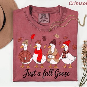 May include: Crimson-colored t-shirt featuring a whimsical illustration of four geese dressed in fall attire. Each goose wears a hat and scarf, with one holding a coffee cup. The text "Just a fall Goose" is printed below the geese.