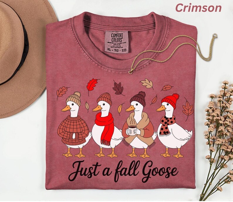 May include: Crimson-colored t-shirt featuring a whimsical illustration of four geese dressed in fall attire. Each goose wears a hat and scarf, with one holding a coffee cup. The text "Just a fall Goose" is printed below the geese.