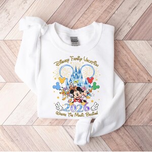 Peut inclure: Sweat-shirt blanc avec un graphisme color&eacute; repr&eacute;sentant des personnages Disney, un ch&acirc;teau et le texte "Disney Family Vacation 2026". Le motif comprend Mickey Mouse, Minnie Mouse, Donald Duck et Dingo. La phrase "Where The Magic Began" est &eacute;galement incluse.