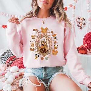 May include: A light pink sweatshirt featuring a castle design within a decorative frame. The design includes fall-themed elements like pumpkins, leaves, and coffee cups. The sweatshirt has long sleeves and a crew neck.
