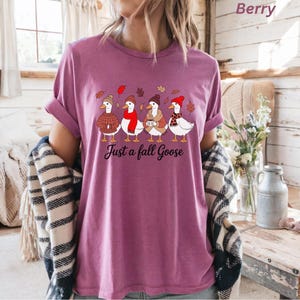 May include: Berry-colored t-shirt featuring a whimsical illustration of five geese wearing hats and scarves, with fall leaves. The text "Just a fall Goose" is printed below the geese. The shirt has rolled-up sleeves.
