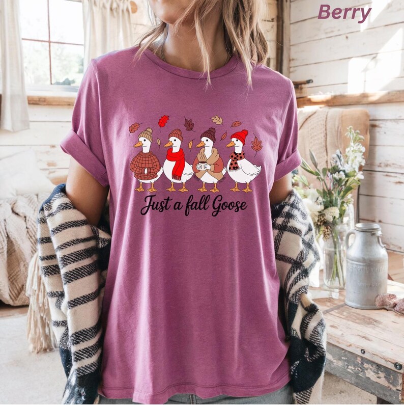 May include: Berry-colored t-shirt featuring a whimsical illustration of five geese wearing hats and scarves, with fall leaves. The text "Just a fall Goose" is printed below the geese. The shirt has rolled-up sleeves.