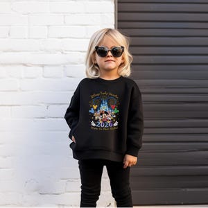 Peut inclure: Un sweat-shirt noir avec un graphisme color&eacute; de Mickey et Minnie Mouse devant un ch&acirc;teau, avec le texte "Disney Family Vacation 2026 Where The Magic Begins". Le sweat-shirt est port&eacute; par un jeune enfant.