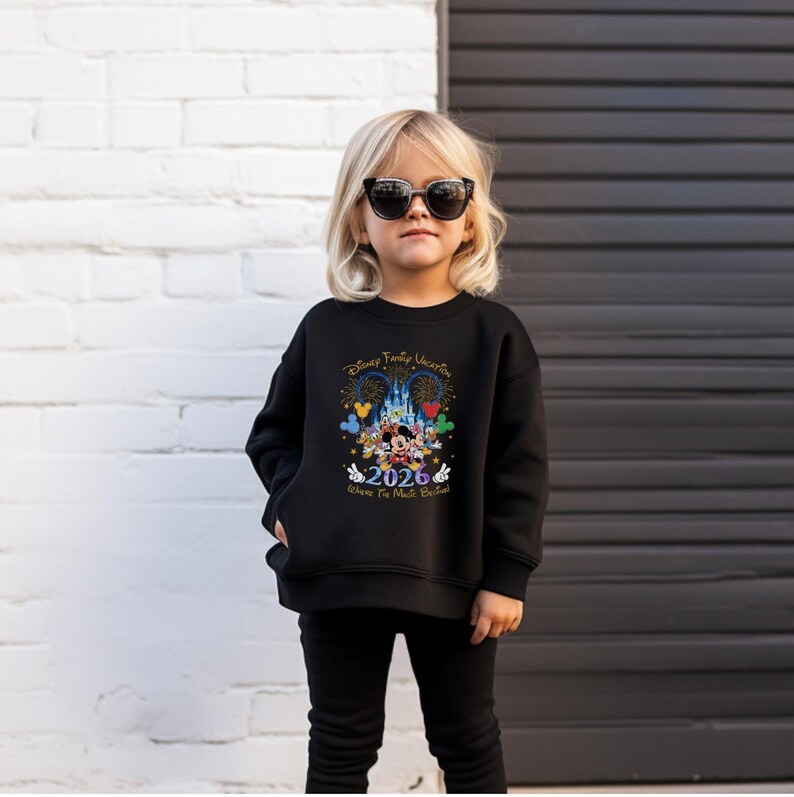 Peut inclure: Un sweat-shirt noir avec un graphisme color&eacute; de Mickey et Minnie Mouse devant un ch&acirc;teau, avec le texte "Disney Family Vacation 2026 Where The Magic Begins". Le sweat-shirt est port&eacute; par un jeune enfant.