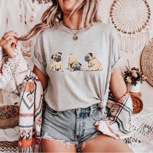 May include: A light gray t-shirt featuring three cartoon pugs in different poses. The pugs are tan with black markings. The shirt is worn with denim shorts and a patterned shawl.