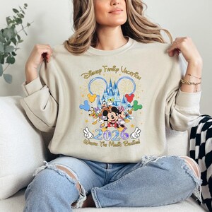 Peut inclure: Sweat-shirt beige avec un motif Disney color&eacute;. Le motif comprend Mickey Mouse, Donald Duck et Dingo, ainsi que le texte "Disney Family Vacation 2026". Le motif comprend un ch&acirc;teau et des feux d'artifice.