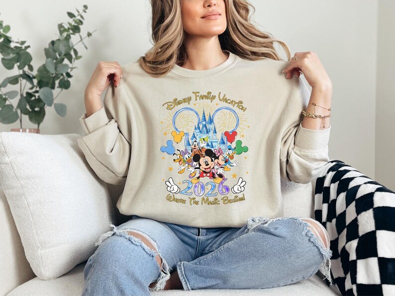 Peut inclure: Sweat-shirt beige avec un motif Disney color&eacute;. Le motif comprend Mickey Mouse, Donald Duck et Dingo, ainsi que le texte "Disney Family Vacation 2026". Le motif comprend un ch&acirc;teau et des feux d'artifice.