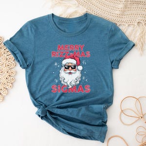May include: A teal blue t-shirt with a graphic of Santa Claus wearing sunglasses and the text "Merry Rizzmas Sigmas".