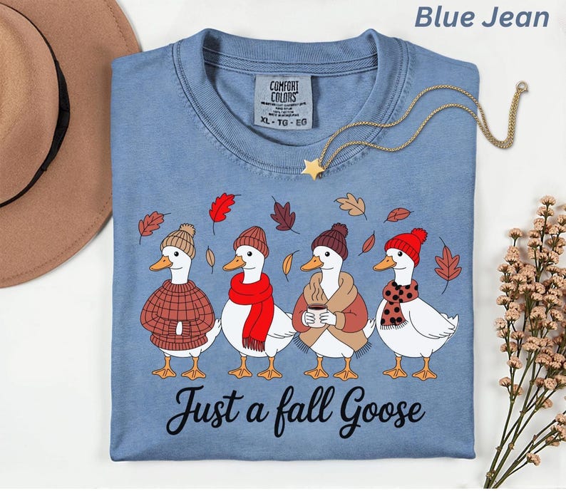 May include: Blue jean colored t-shirt with a graphic of four geese wearing fall attire. The geese are wearing hats, scarves, and sweaters. The text "Just a fall Goose" is printed below the geese. A gold necklace with a star pendant is on the shirt.