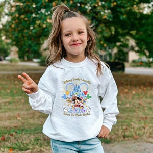 Peut inclure: Un sweat-shirt blanc &agrave; manches longues avec un graphisme color&eacute; de personnages Disney, un ch&acirc;teau et le texte "Disney Family Vacation 2026". Le sweat-shirt est port&eacute; par une jeune personne.