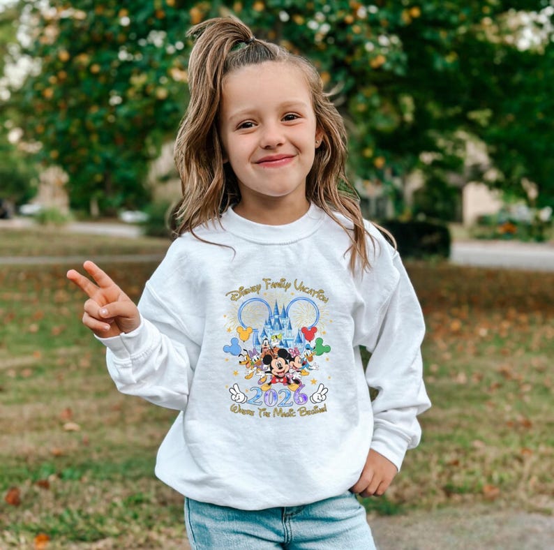 Peut inclure: Un sweat-shirt blanc &agrave; manches longues avec un graphisme color&eacute; de personnages Disney, un ch&acirc;teau et le texte "Disney Family Vacation 2026". Le sweat-shirt est port&eacute; par une jeune personne.