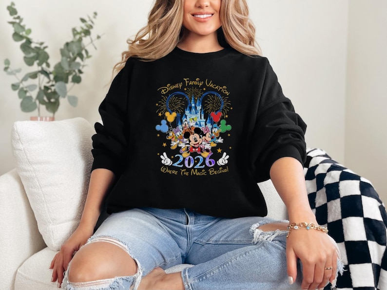 Peut inclure: Sweat-shirt noir avec un motif de vacances en famille Disney. Le graphisme comprend Mickey, Minnie, Donald et Dingo devant un ch&acirc;teau, avec l'ann&eacute;e 2026 et la phrase "Where the Magic Begins!"