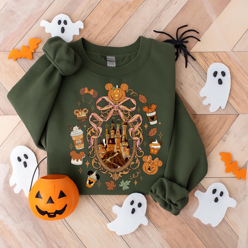 May include: An olive green sweatshirt featuring a Halloween-themed graphic with a castle, pumpkins, and Mickey Mouse ears. The design is surrounded by fall-themed elements. Decorative ghosts, bats, and a jack-o'-lantern surround the sweatshirt.
