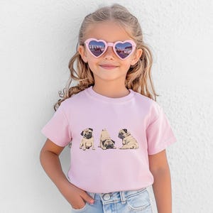 May include: A young girl wearing heart-shaped sunglasses and a light pink t-shirt. The t-shirt features a graphic of three pugs in different poses. The girl is wearing blue jeans.