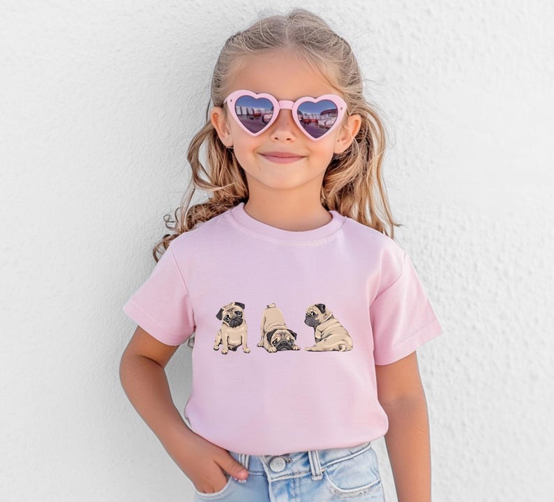 May include: A young girl wearing heart-shaped sunglasses and a light pink t-shirt. The t-shirt features a graphic of three pugs in different poses. The girl is wearing blue jeans.