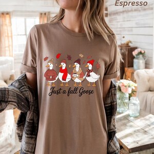 May include: Espresso-colored t-shirt featuring a whimsical illustration of four geese dressed in fall attire, including hats, scarves, and sweaters. The text "Just a fall Goose" is printed below the geese.