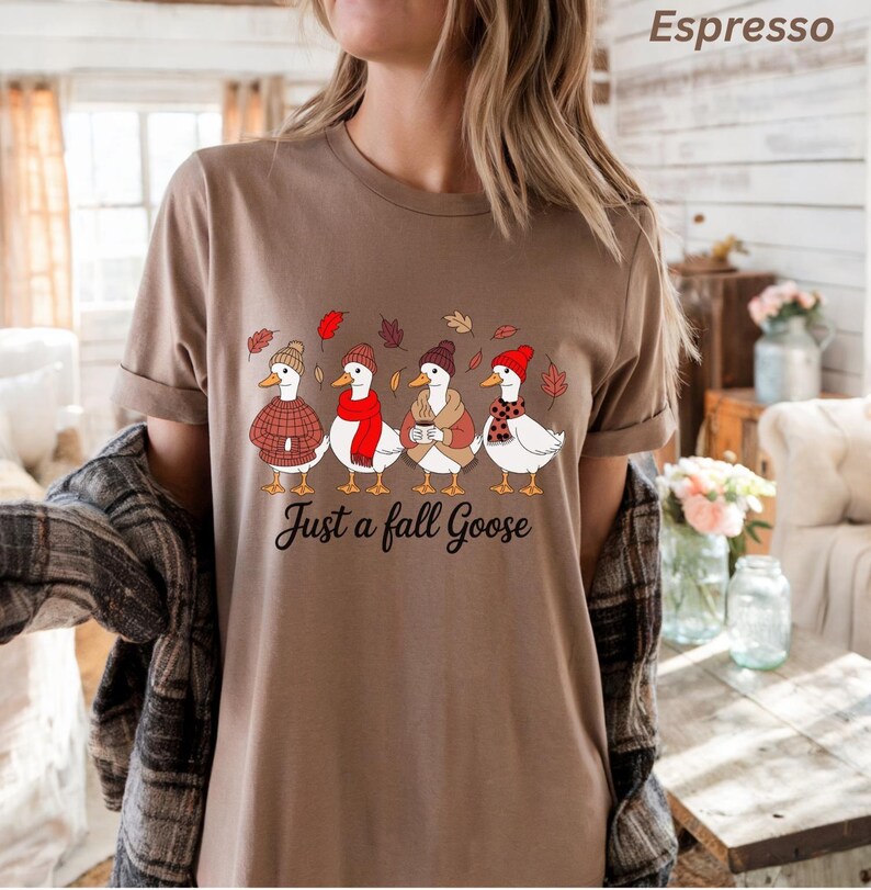 May include: Espresso-colored t-shirt featuring a whimsical illustration of four geese dressed in fall attire, including hats, scarves, and sweaters. The text "Just a fall Goose" is printed below the geese.