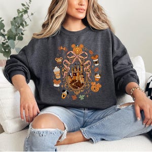 May include: Dark gray sweatshirt featuring a colorful graphic design. The design includes a castle, pumpkins, coffee cups, and Mickey Mouse. The sweatshirt has long sleeves and a crew neck. The overall theme is autumn and Disney.