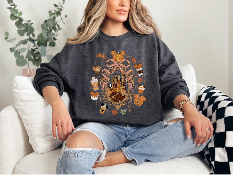 May include: Dark gray sweatshirt featuring a colorful graphic design. The design includes a castle, pumpkins, coffee cups, and Mickey Mouse. The sweatshirt has long sleeves and a crew neck. The overall theme is autumn and Disney.