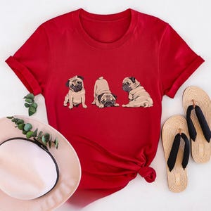 May include: A red t-shirt featuring three cartoon pugs in different poses. The shirt is paired with a sun hat and two pairs of beige flip-flops with black straps. The shirt is a vibrant red color.