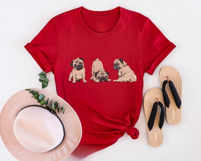 May include: A red t-shirt featuring three cartoon pugs in different poses. The shirt is paired with a sun hat and two pairs of beige flip-flops with black straps. The shirt is a vibrant red color.