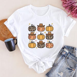 Puede incluir: Camiseta blanca con un gr&aacute;fico de varias calabazas con sombreros de vaquero. Las calabazas son de color naranja y marr&oacute;n con un dise&ntilde;o de estampado de leopardo.